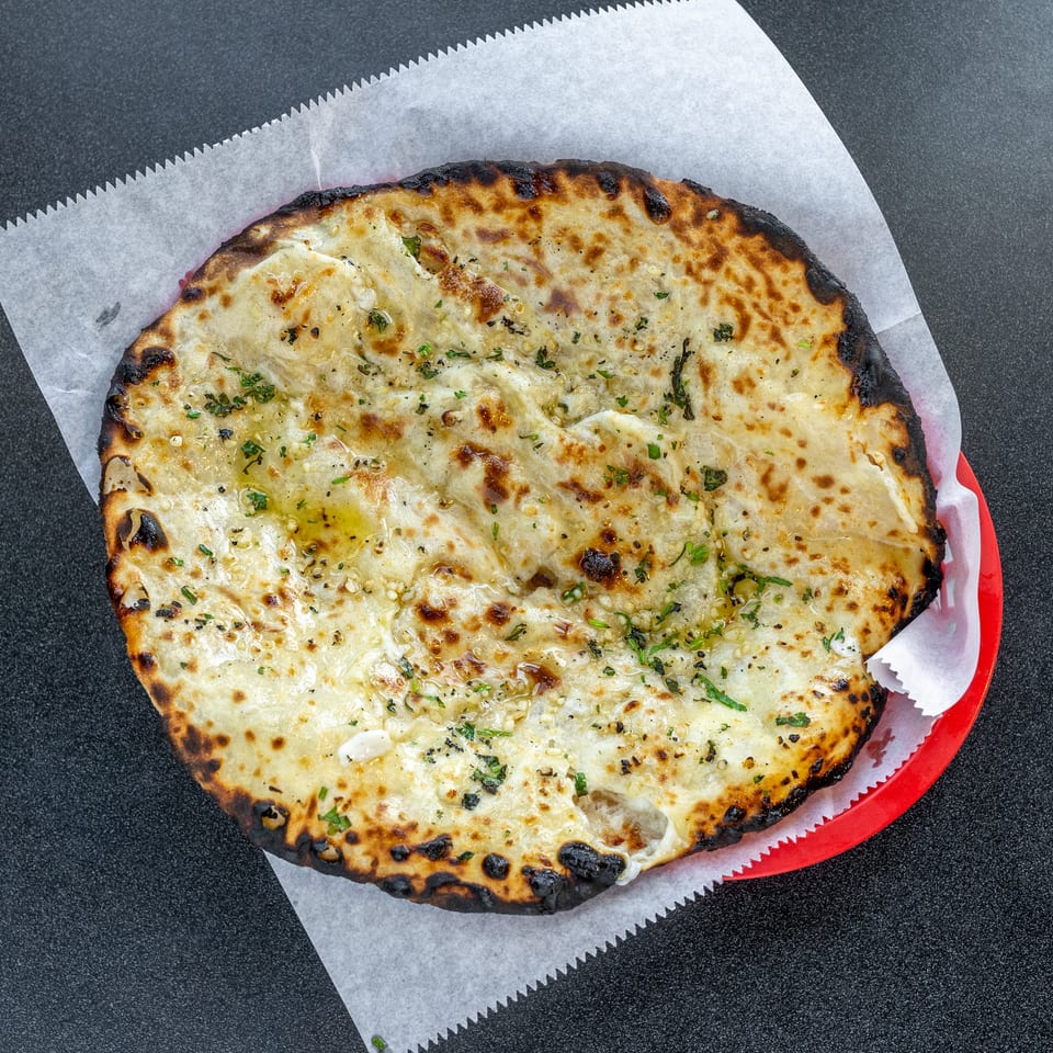 Large blistered flatbread topped with melted cheese, herbs, garlic, and oil, served on parchment in a red tray.
