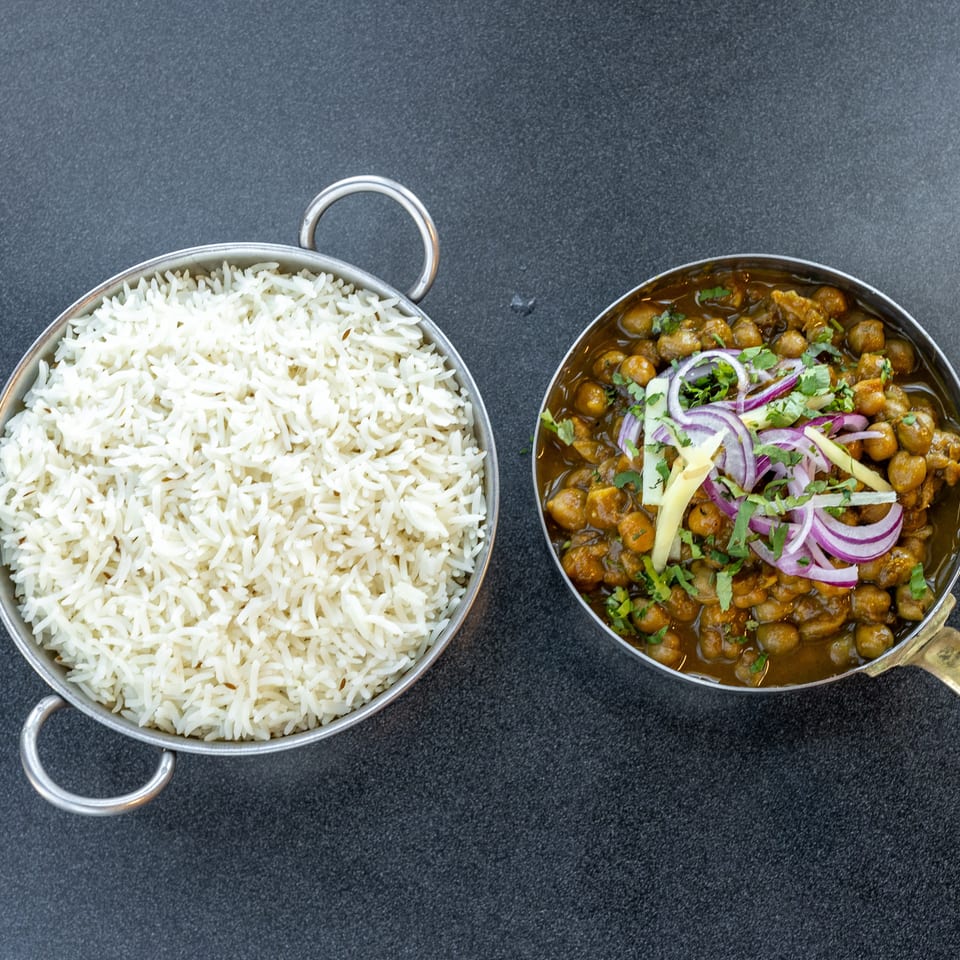 Overhead view of fragrant white rice beside chickpea curry garnished with red onions, ginger, and cilantro in metal serving pans.