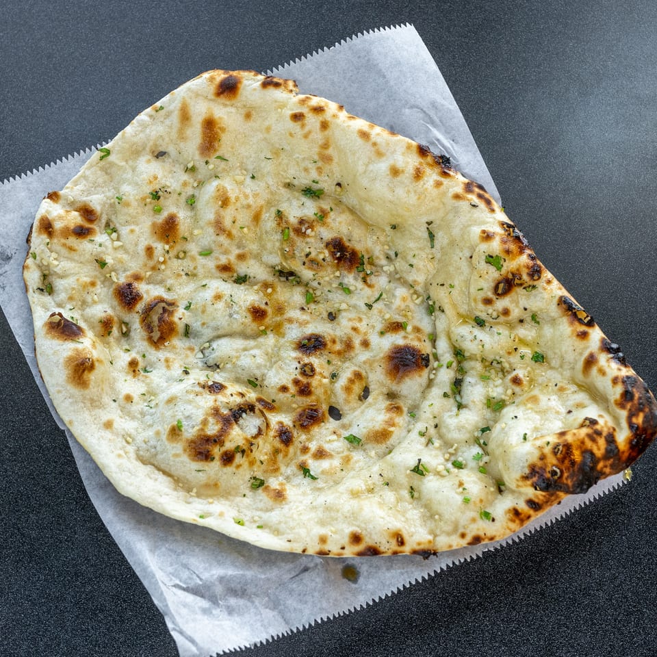 Large garlic naan with charred spots and chopped herbs served on parchment paper.