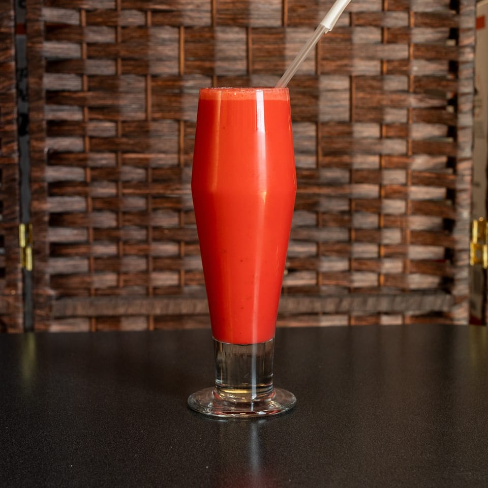 Bright red blended drink in a tall glass with a straw, displayed on a dark table against a rustic wooden wall.