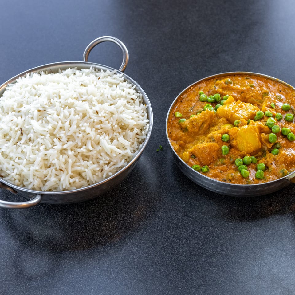 Metal serving dishes hold fluffy white rice and creamy orange curry with potatoes and green peas on a dark tabletop.