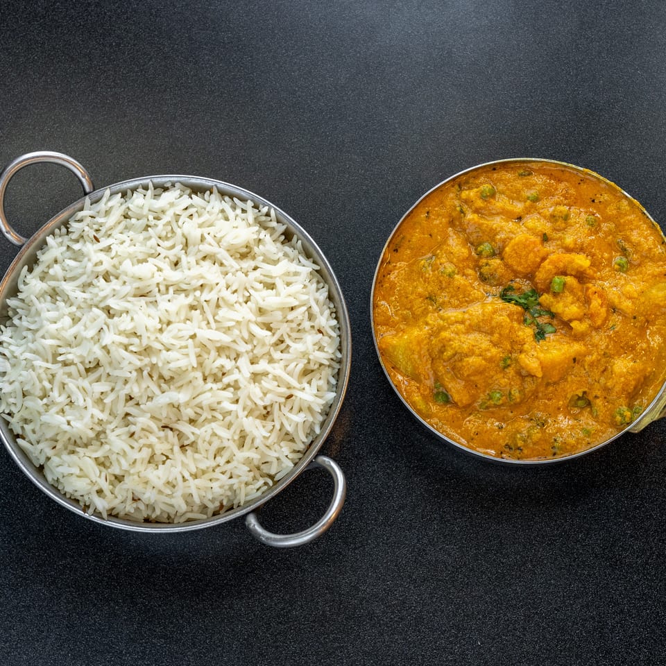 Basmati rice served beside creamy orange curry with vegetables in metal pans on a dark tabletop.