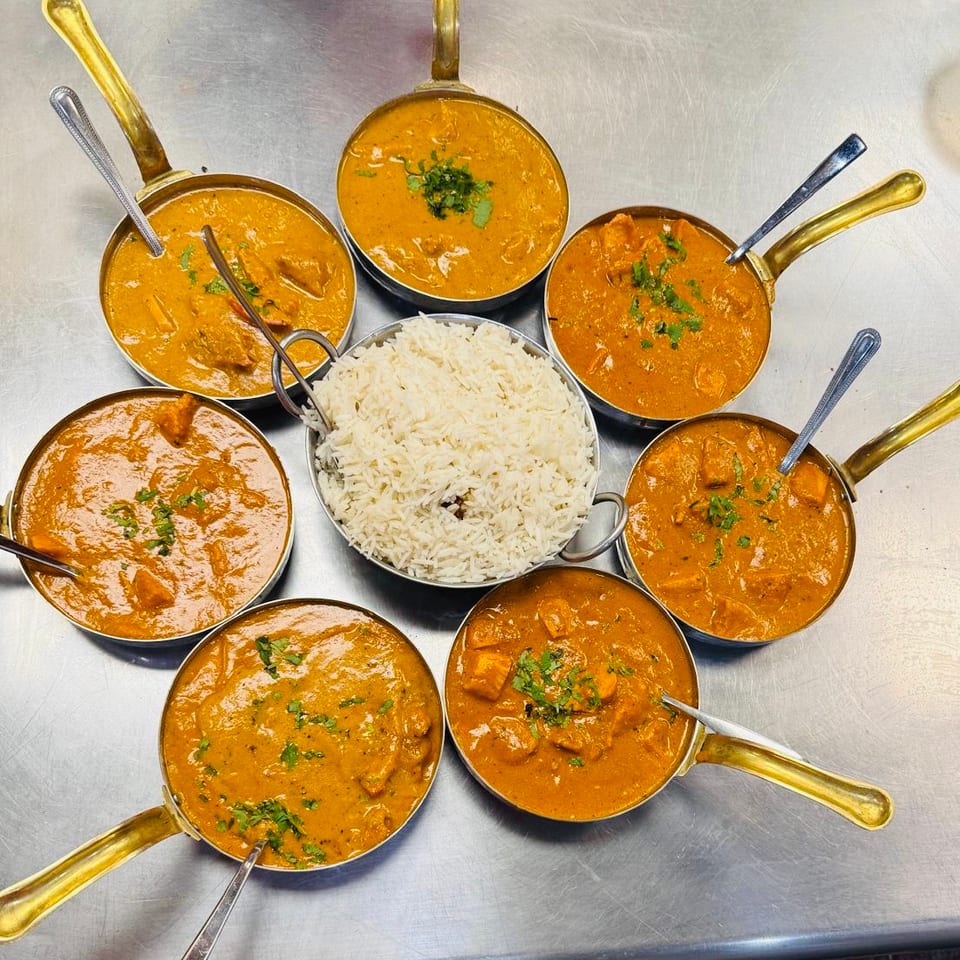 Overhead view of seven bowls of creamy orange Indian curries arranged around a central bowl of basmati rice on a stainless-steel counter.