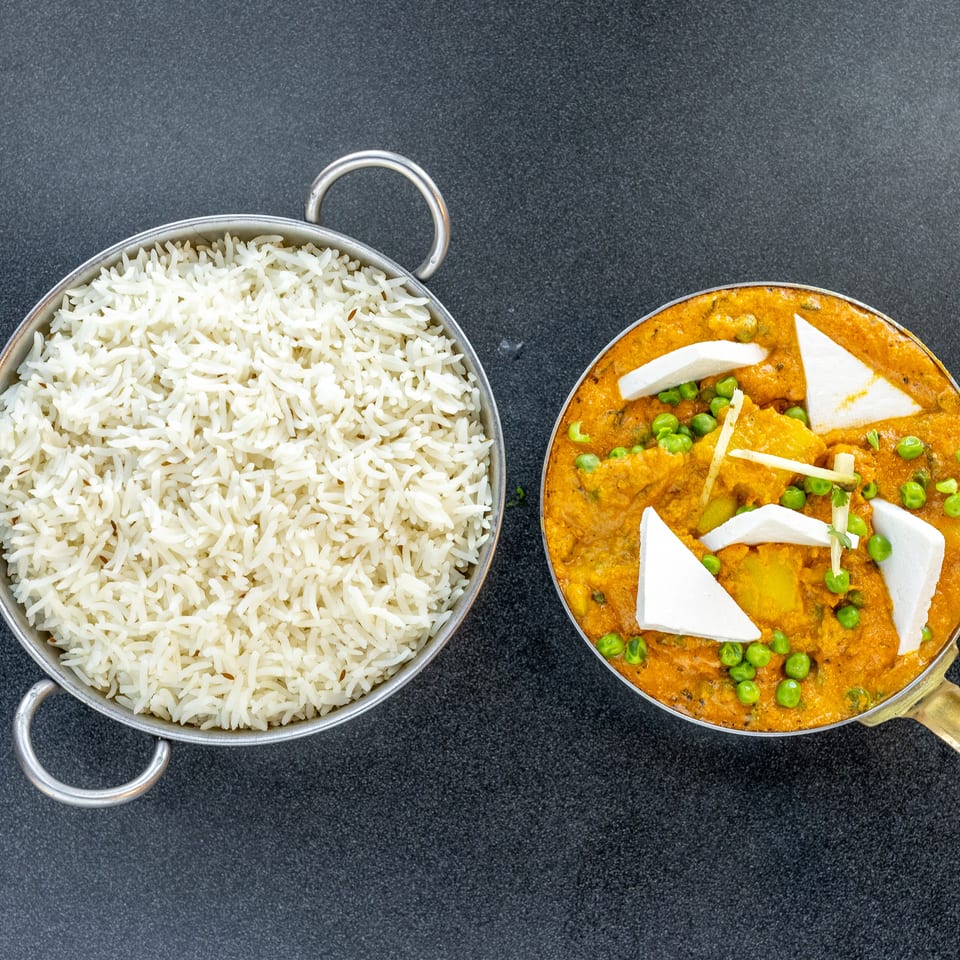 Overhead view of basmati rice beside creamy orange curry with peas and paneer, served in metal pans on a dark surface.