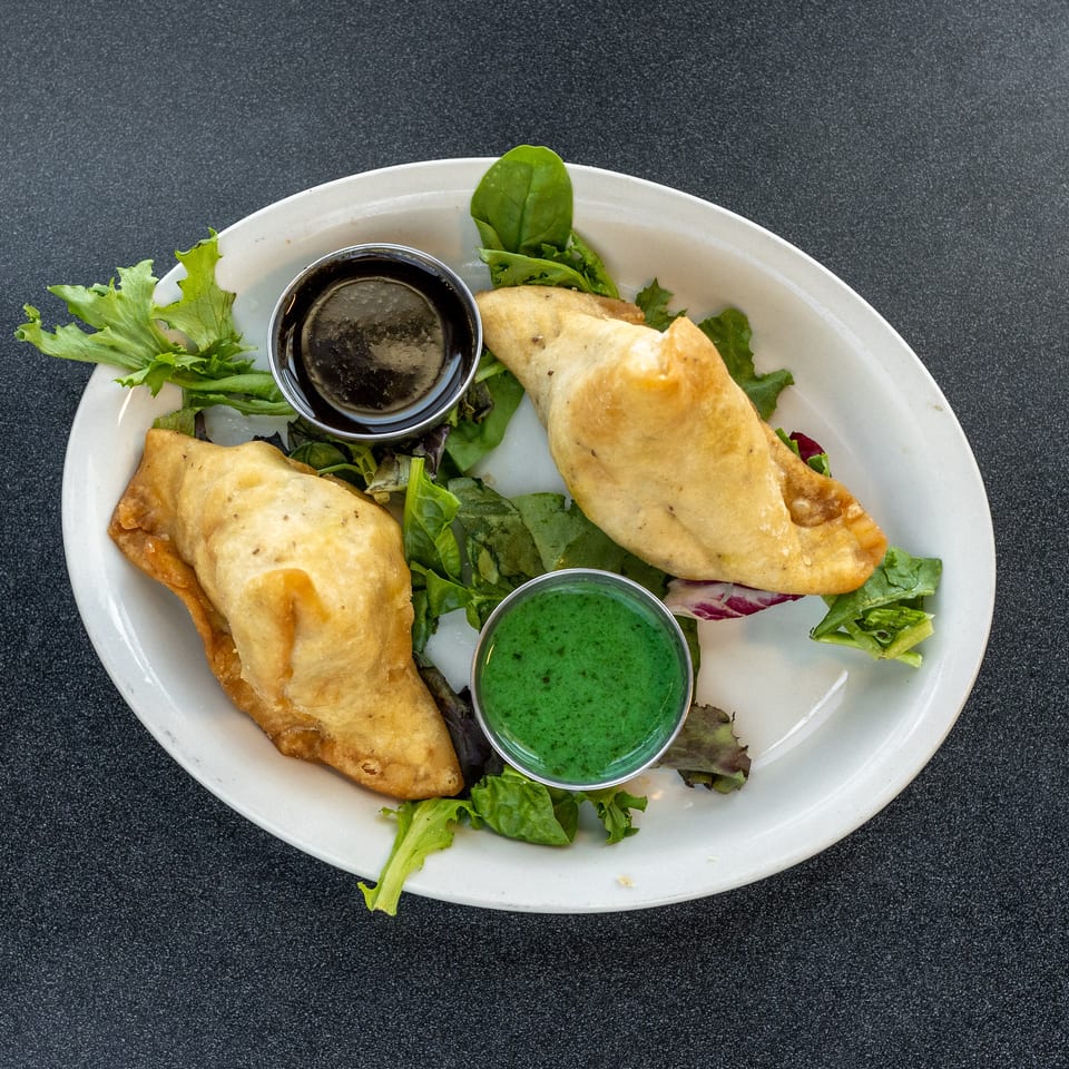 Plate of golden fried turnovers on mixed greens with small cups of dark and green dipping sauces.